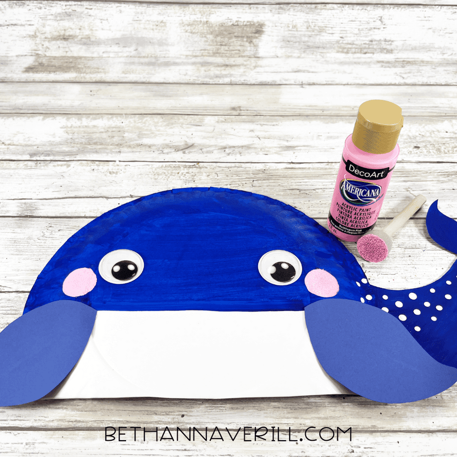 whale craft with painted cheeks, dotted tail details, and googly eyes, with pink paint and a sponge dabber beside it.