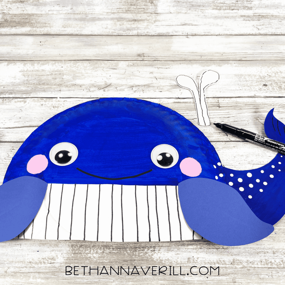 Whale paper plate craft with drawn vertical lines on the belly and a cut-out paper water spout placed above the head, with a black marker beside it.