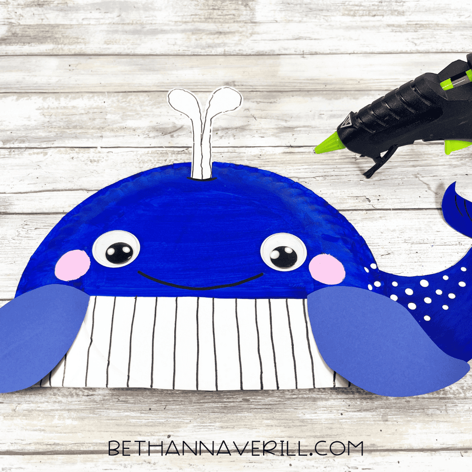 Whale paper plate craft with the water spout glued onto the top of the head, and a glue gun positioned to the side.