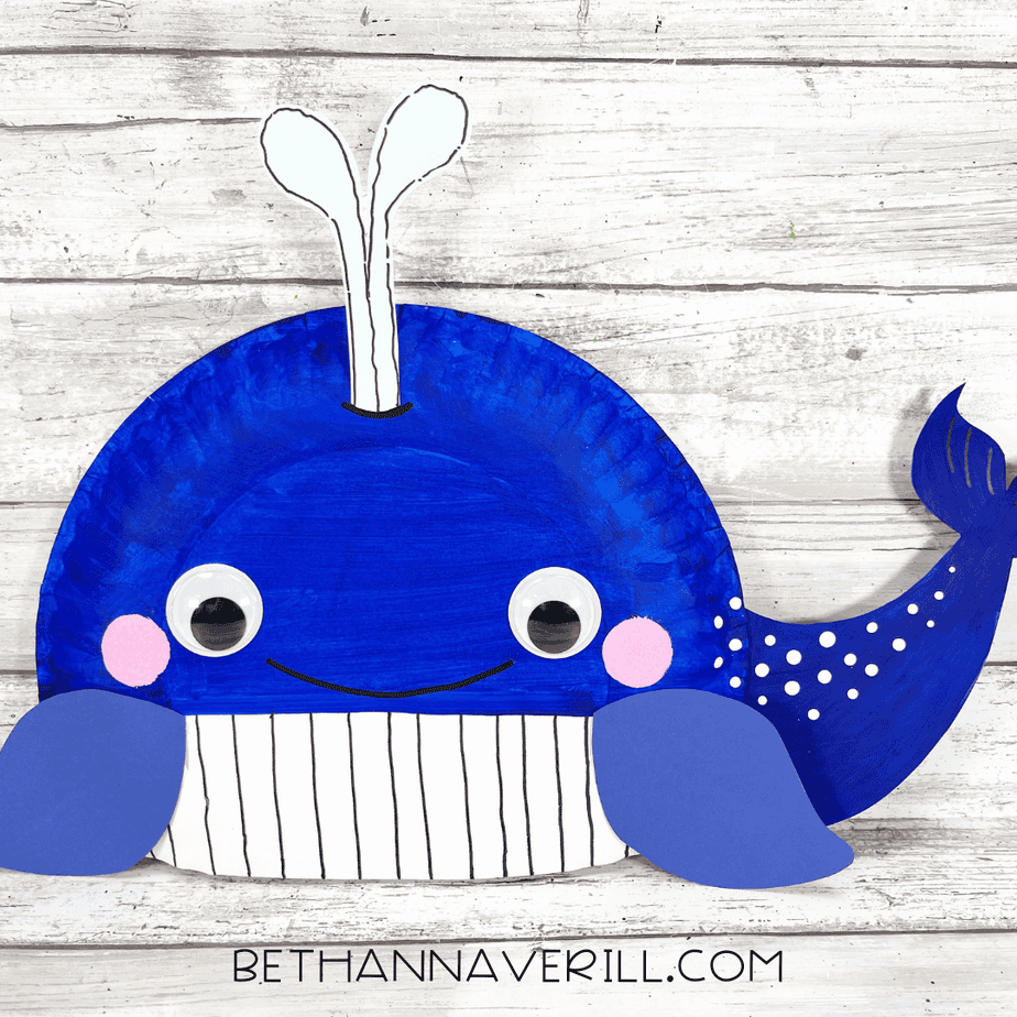 Finished blue whale paper plate craft with googly eyes, pink cheeks, dotted tail, striped belly, and a white paper water spout attached to the top.
