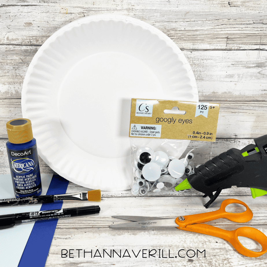 Supplies needed for a whale paper plate craft including a white paper plate, blue acrylic paint, googly eyes, glue gun, scissors, paintbrush, black marker, and blue cardstock arranged on a wooden background.