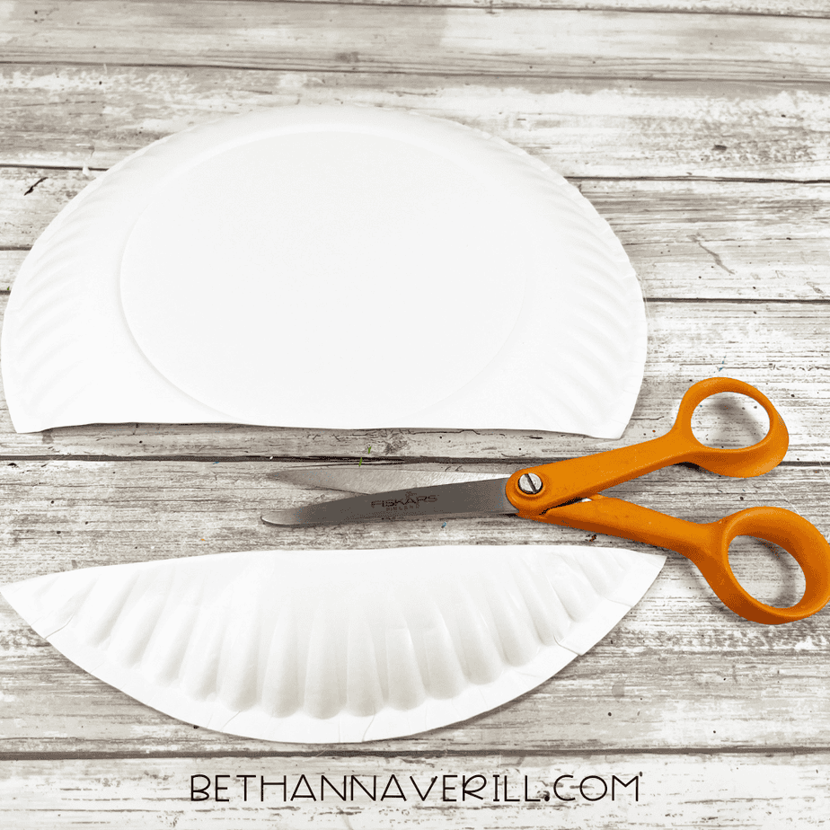 A white paper plate cut in half with scissors placed beside it for the start of a whale paper plate craft.