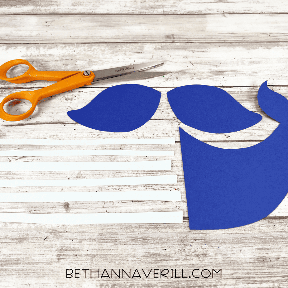 Cut-out blue cardstock whale tail and fins laid out with thin white paper strips and scissors on a wooden surface.