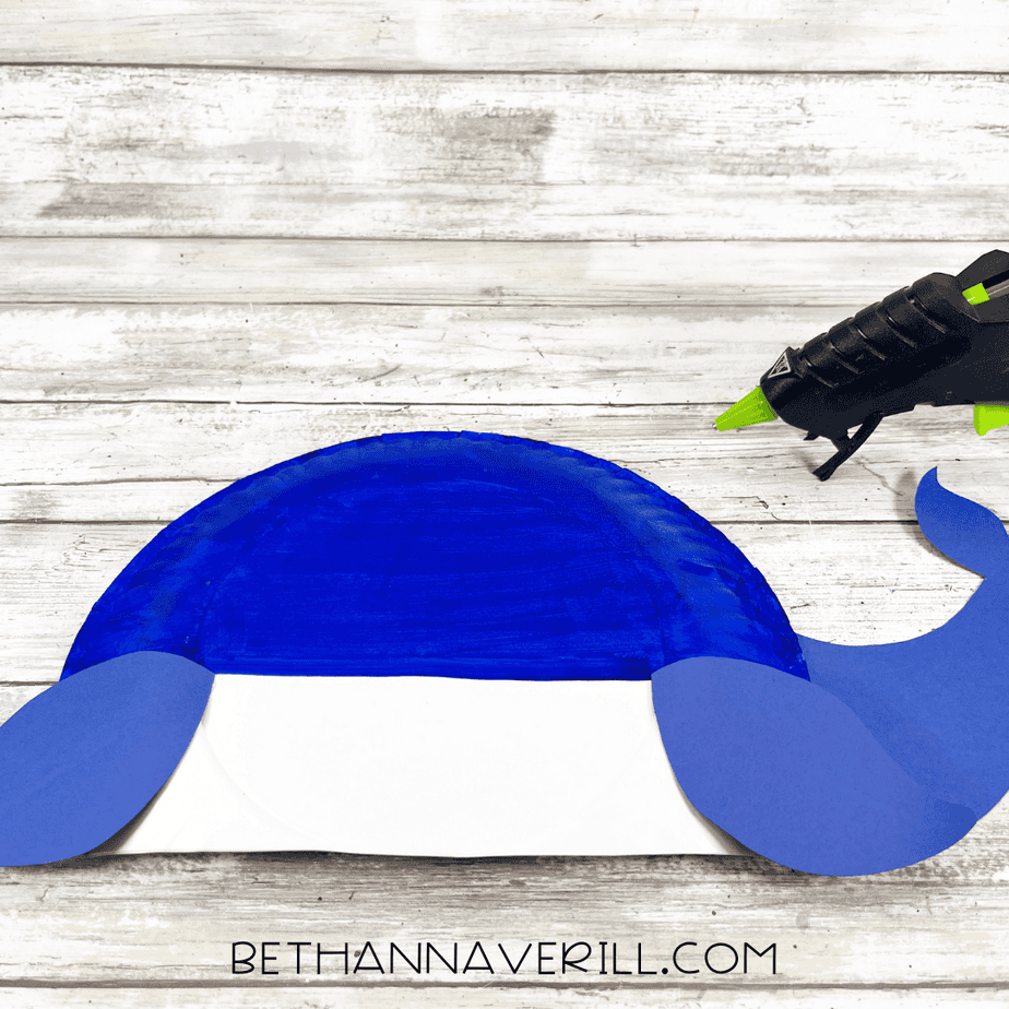 Blue whale body made from a painted paper plate with blue cardstock fins attached, and a glue gun positioned nearby.