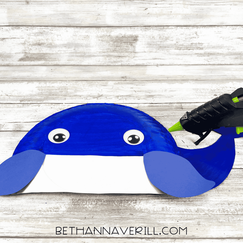Whale craft with googly eyes attached to the painted paper plate body, with a glue gun positioned to the side.