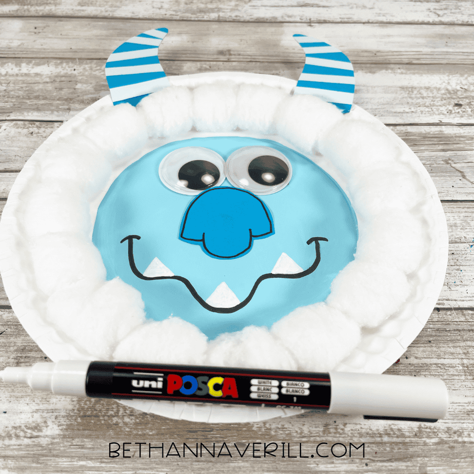 Yeti paper plate craft with googly eyes, blue nose, striped horns, cotton ball fur, and white teeth, with a white paint pen placed in front.