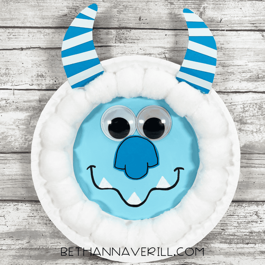 Finished blue yeti paper plate craft with cotton ball fur, striped blue horns, large googly eyes, a blue cardstock nose, and a drawn-on smile with white cardstock teeth, displayed on a wooden background.