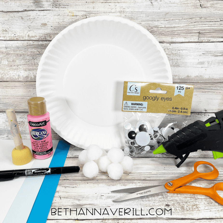 Supplies for a yeti paper plate craft including a white paper plate, pink acrylic paint, sponge dabber, googly eyes, cotton balls, glue gun, scissors, black marker, and blue and white cardstock arranged on a wooden surface.