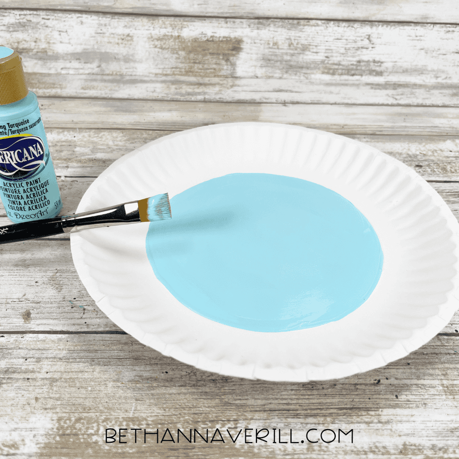 A white paper plate with the center painted light blue using a flat paintbrush, with the bottle of light blue acrylic paint beside it.