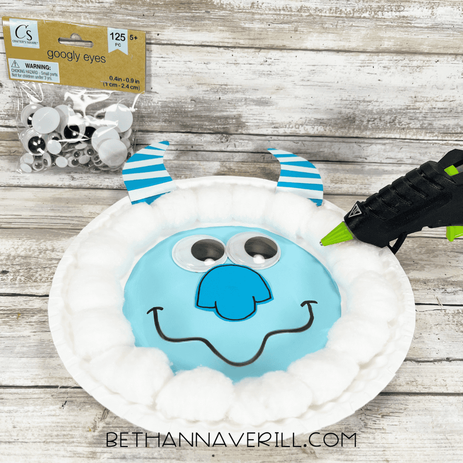 Googly eyes and striped blue horns being glued onto the yeti face on the paper plate using a hot glue gun.