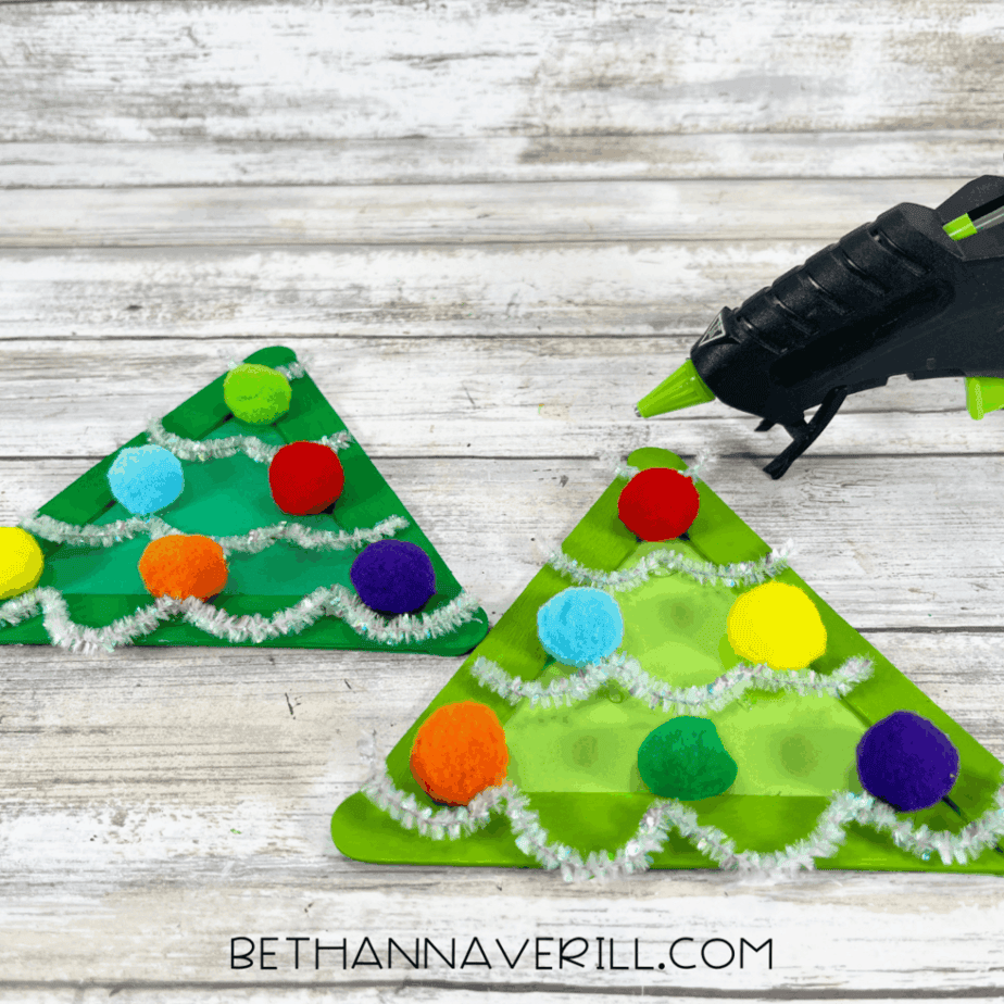 Two decorated green triangle crafts with pom poms and silver pipe cleaner garland, with a hot glue gun positioned nearby.
