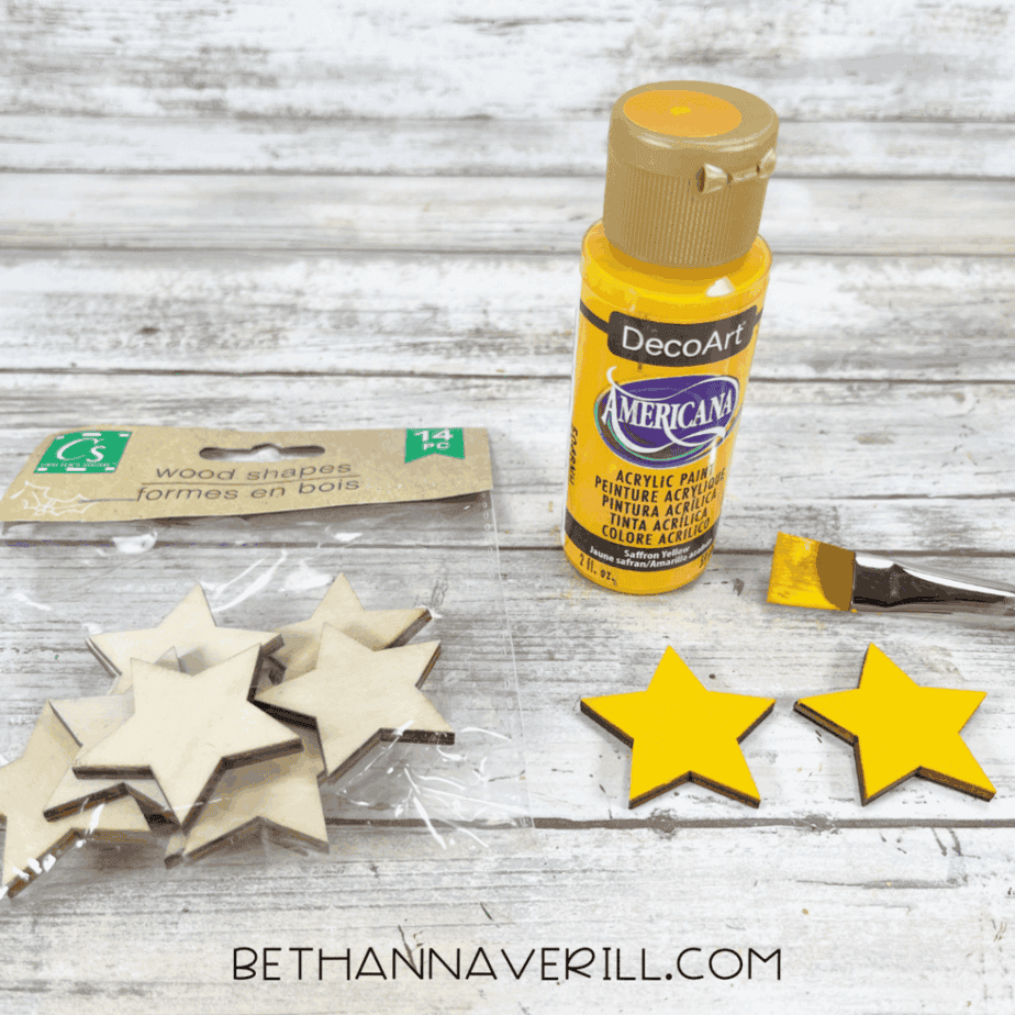 Wooden star shapes and yellow acrylic paint on a white wood surface, with two stars painted yellow and a paintbrush beside them.