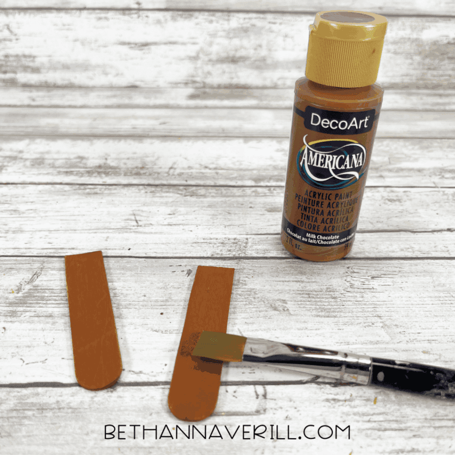 Brown acrylic paint bottle, paintbrush, and two painted popsicle sticks laid on a white wood surface, showing the tree trunk pieces being painted.