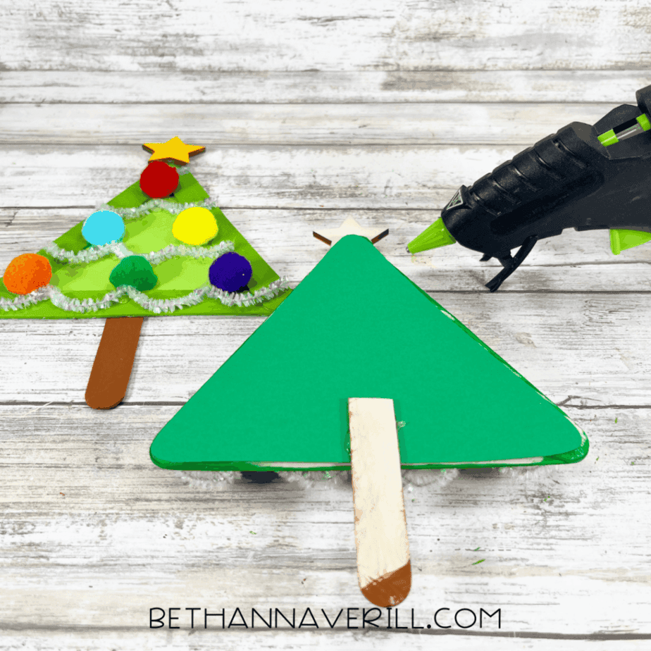 Green cardstock triangle with a popsicle stick attached at the bottom, hot glue gun nearby, and a completed decorated tree visible in the background.