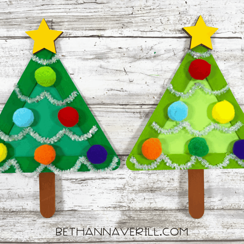 Two completed popsicle stick Christmas tree crafts side by side on a white wood surface, each decorated with pom poms, silver pipe cleaner garland, yellow stars, and brown popsicle stick trunks.