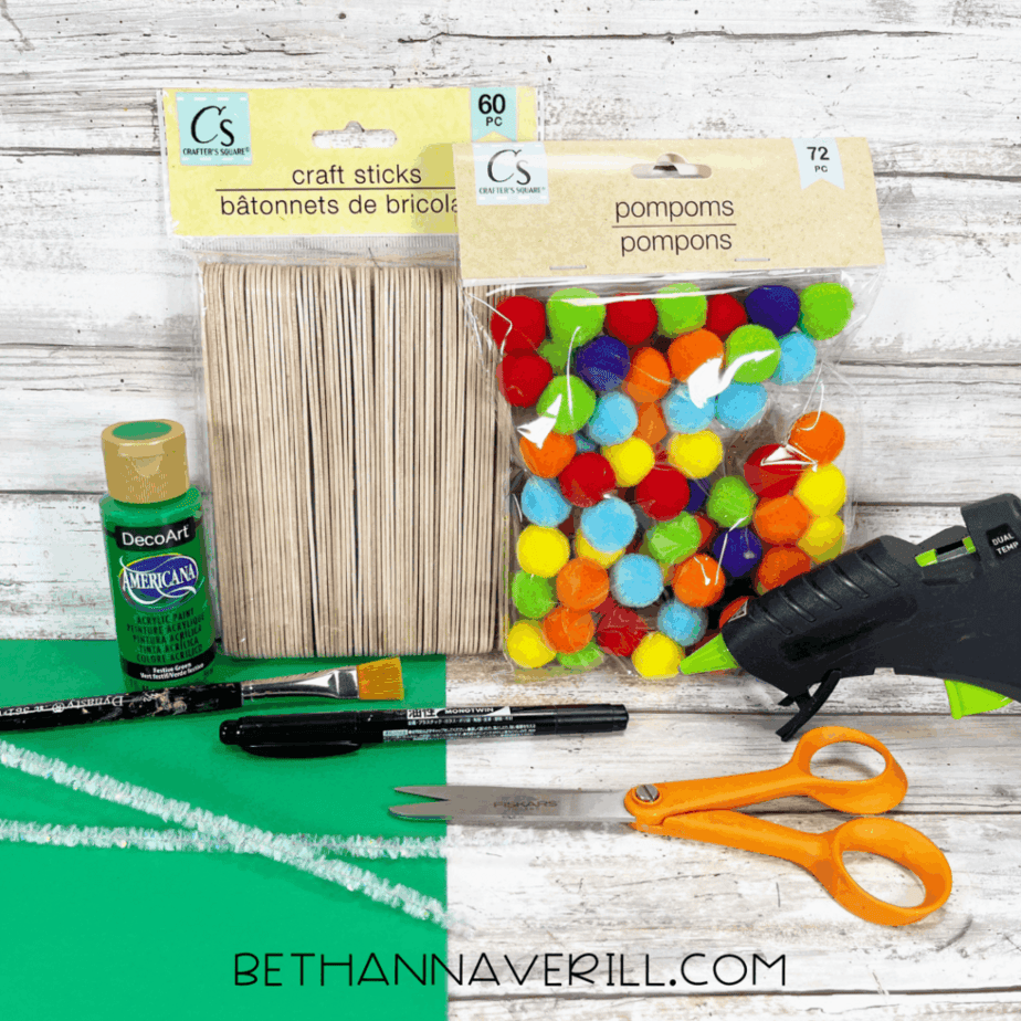 Craft supplies for a Christmas tree popsicle stick craft laid out on a white wood surface, including craft sticks, pom poms, green cardstock, green acrylic paint, pipe cleaners, scissors, paintbrush, marker, and hot glue gun.