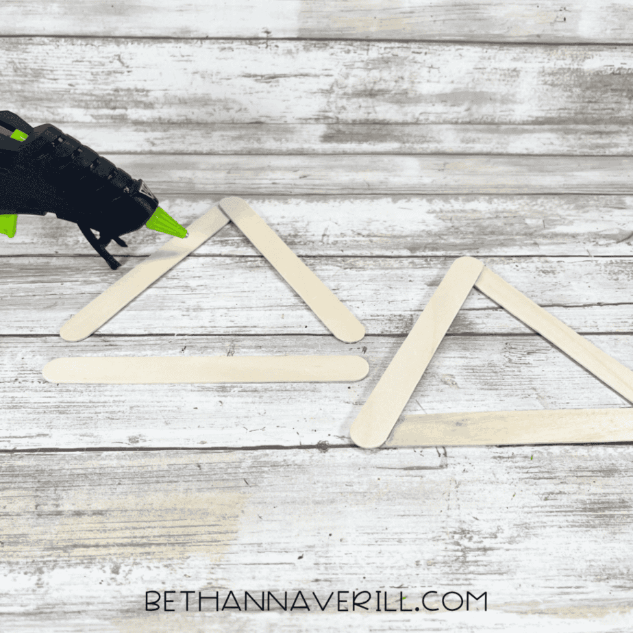 Plain wooden craft sticks arranged into triangle shapes on a white wood surface, with a hot glue gun nearby.