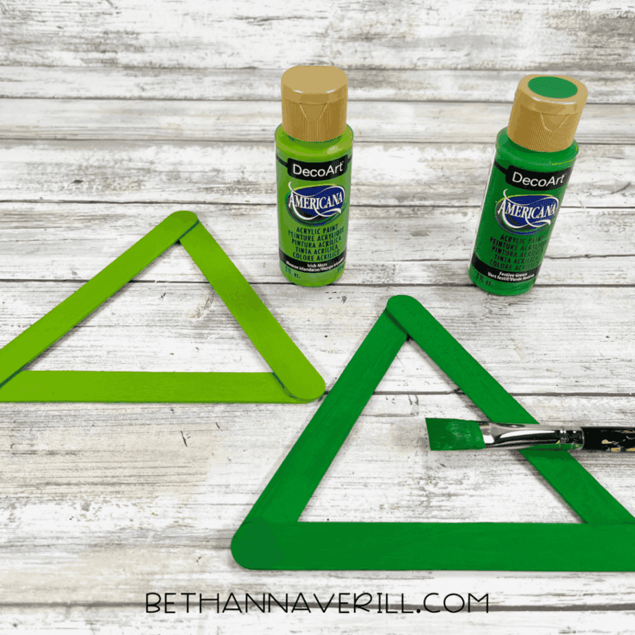 Triangle shapes made from craft sticks painted in two different shades of green, with green acrylic paint bottles and a paintbrush on the table.