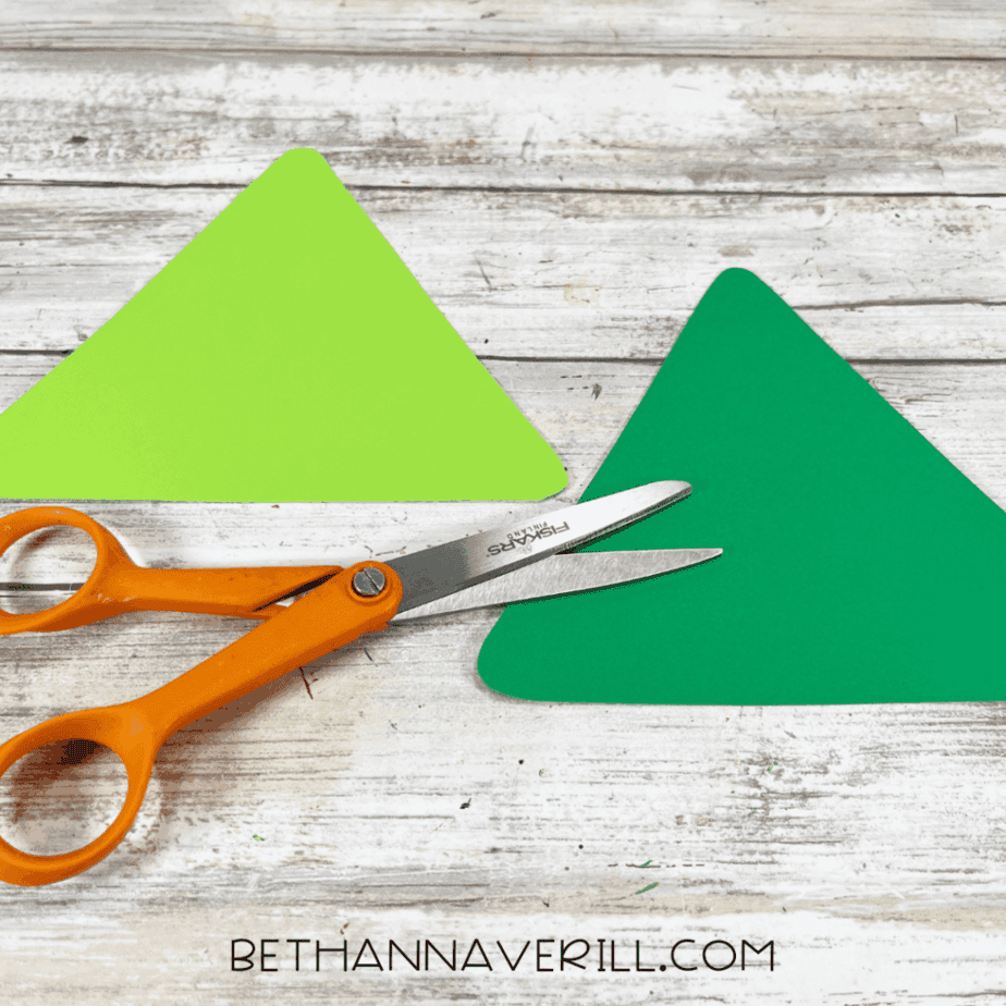 Cut cardstock triangles in two shades of green on a white wood surface, with orange-handled scissors nearby.