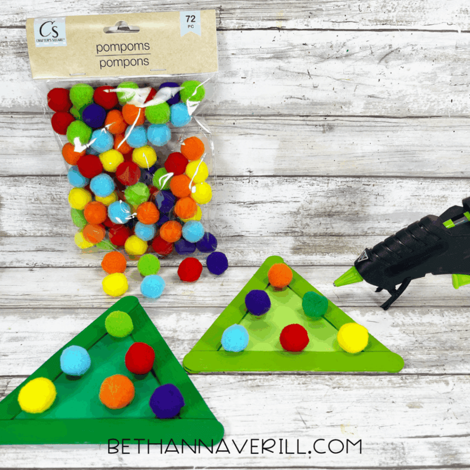 Pom poms being glued onto green triangle shapes made from popsicle sticks and cardstock, with a bag of pom poms and a hot glue gun visible.