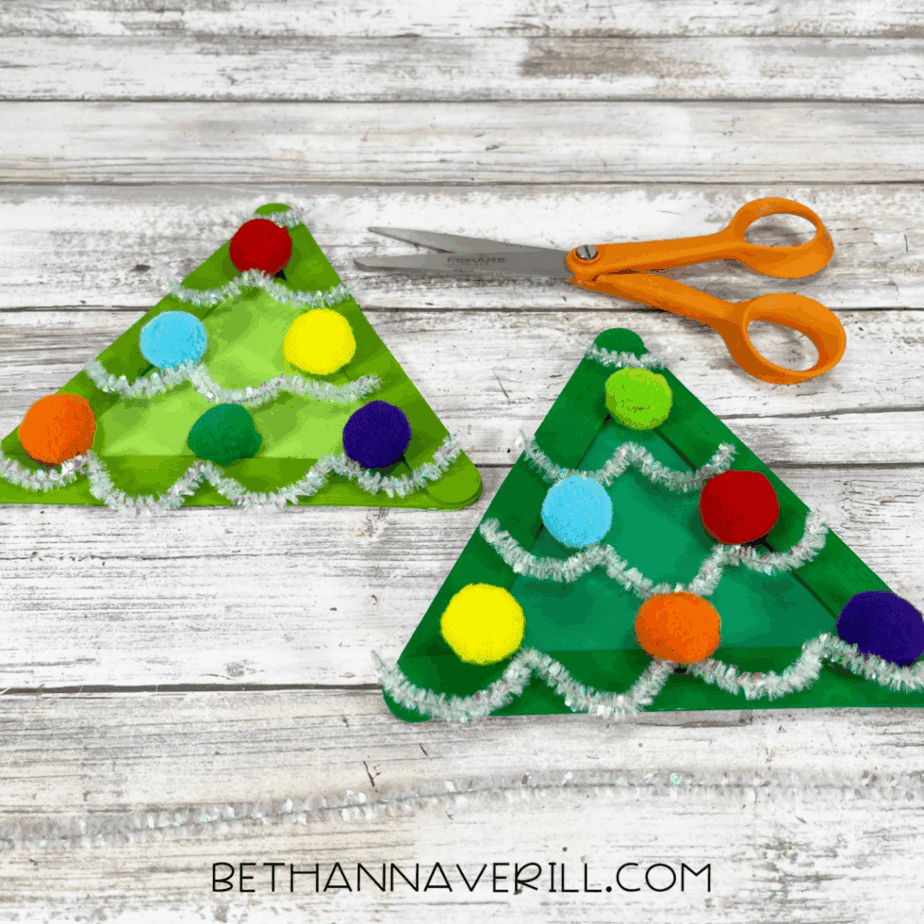 Green triangle crafts decorated with pom poms and silver pipe cleaners arranged like garland, with scissors and extra pipe cleaners on the table.
