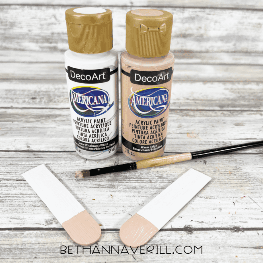 Two acrylic paint bottles labeled white and warm beige with a paintbrush and partially painted craft sticks on a white wood background.