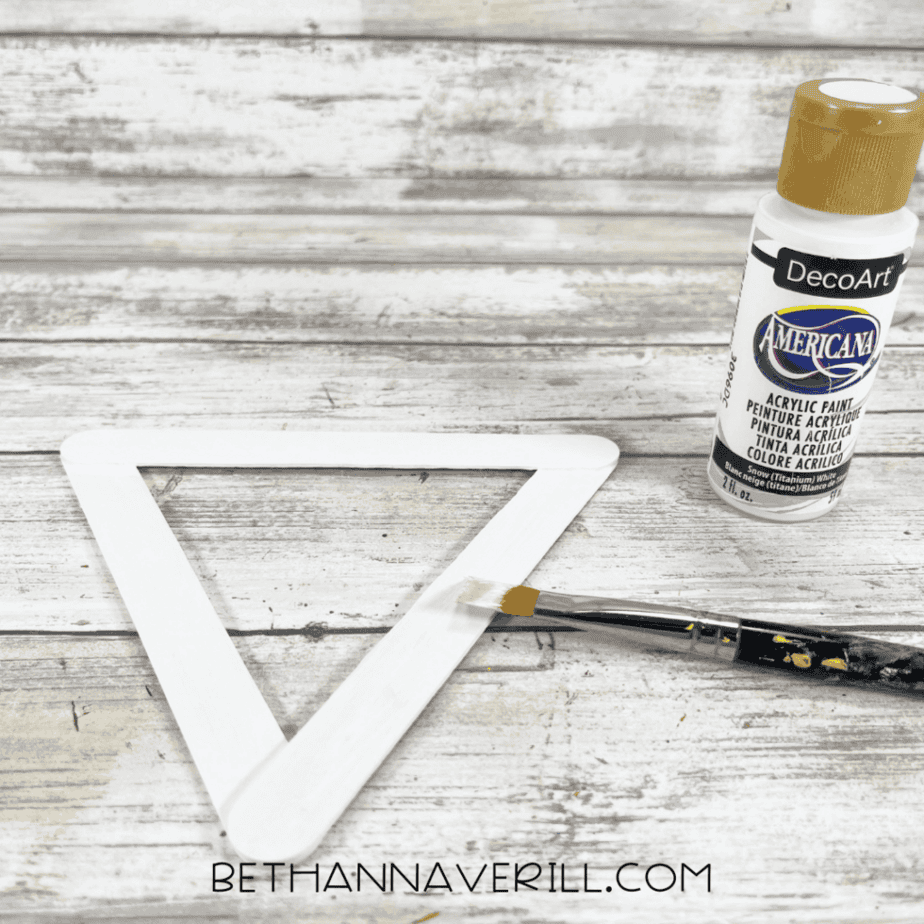 Three glued craft sticks painted white forming a triangular shape, with a paintbrush and white acrylic paint bottle nearby.
