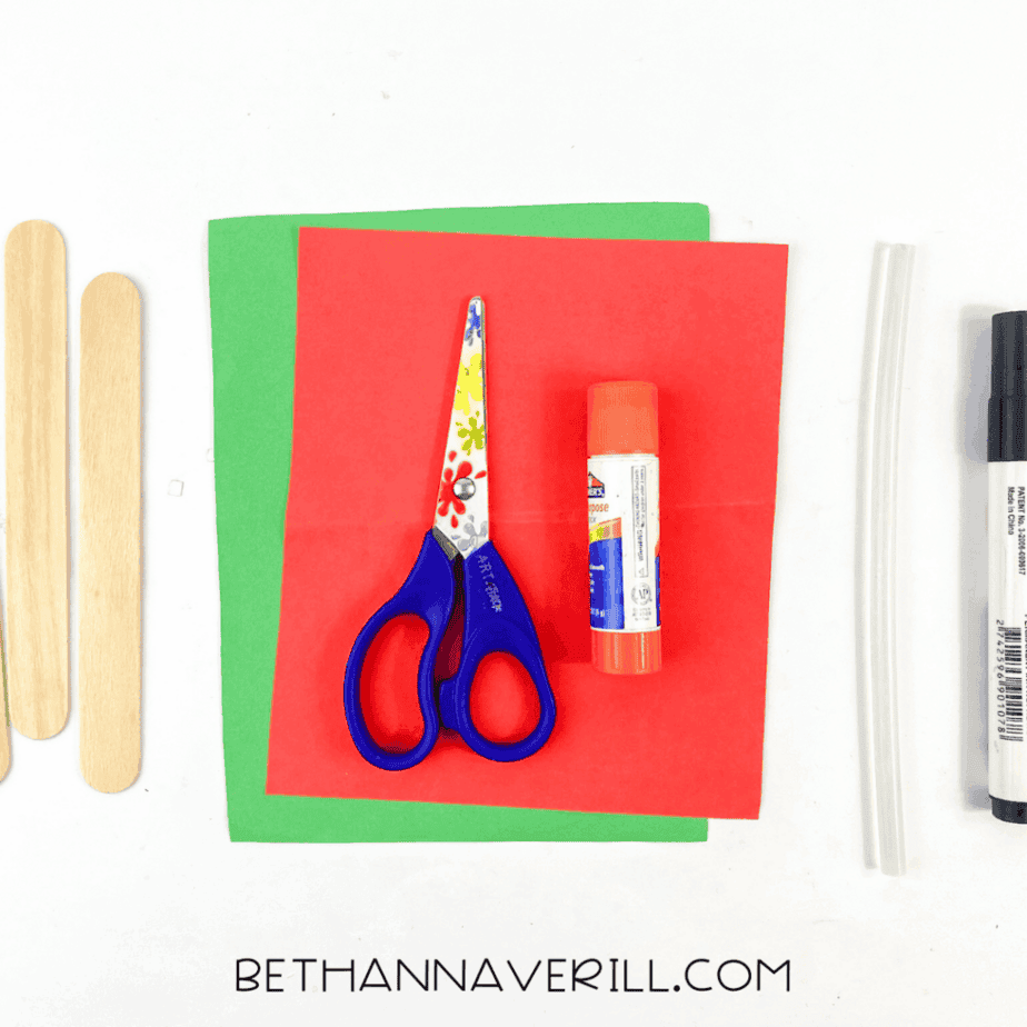 Craft supplies arranged on a white background including jumbo craft sticks, red and green cardstock, scissors, glue stick, glue gun sticks, and black marker.