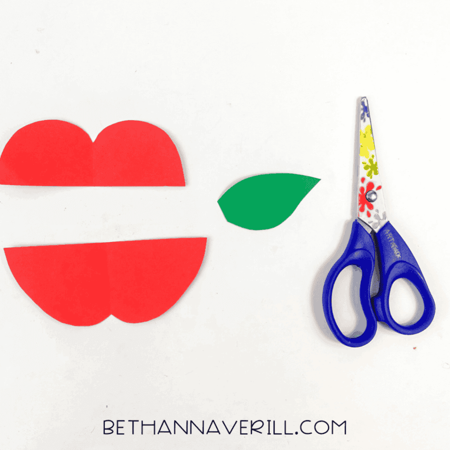 Apple shape pieces cut from red cardstock with a green leaf beside scissors on a white surface.