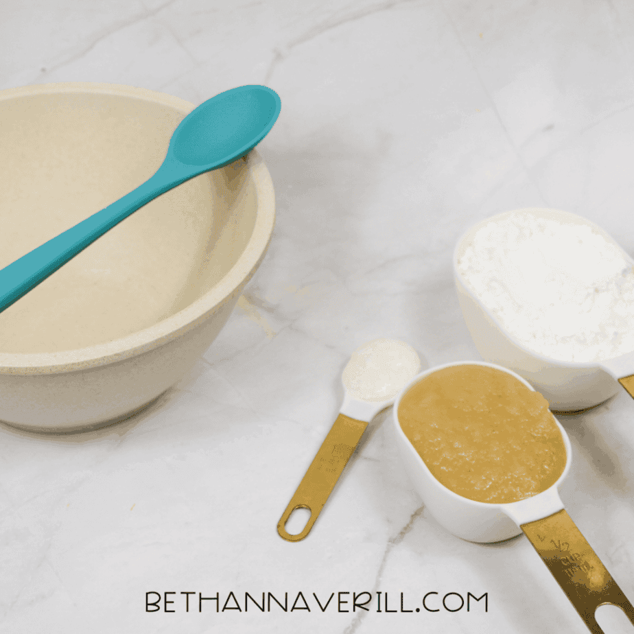 Mixing bowl with teal spoon beside measured applesauce, cornstarch, and liquid on a white surface.