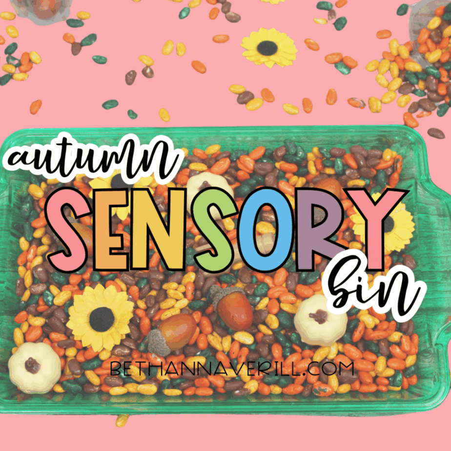 Autumn sensory bin filled with dyed beans, small pumpkins, acorns, and sunflower decorations on a pink background with text reading “Autumn Sensory Bin.”