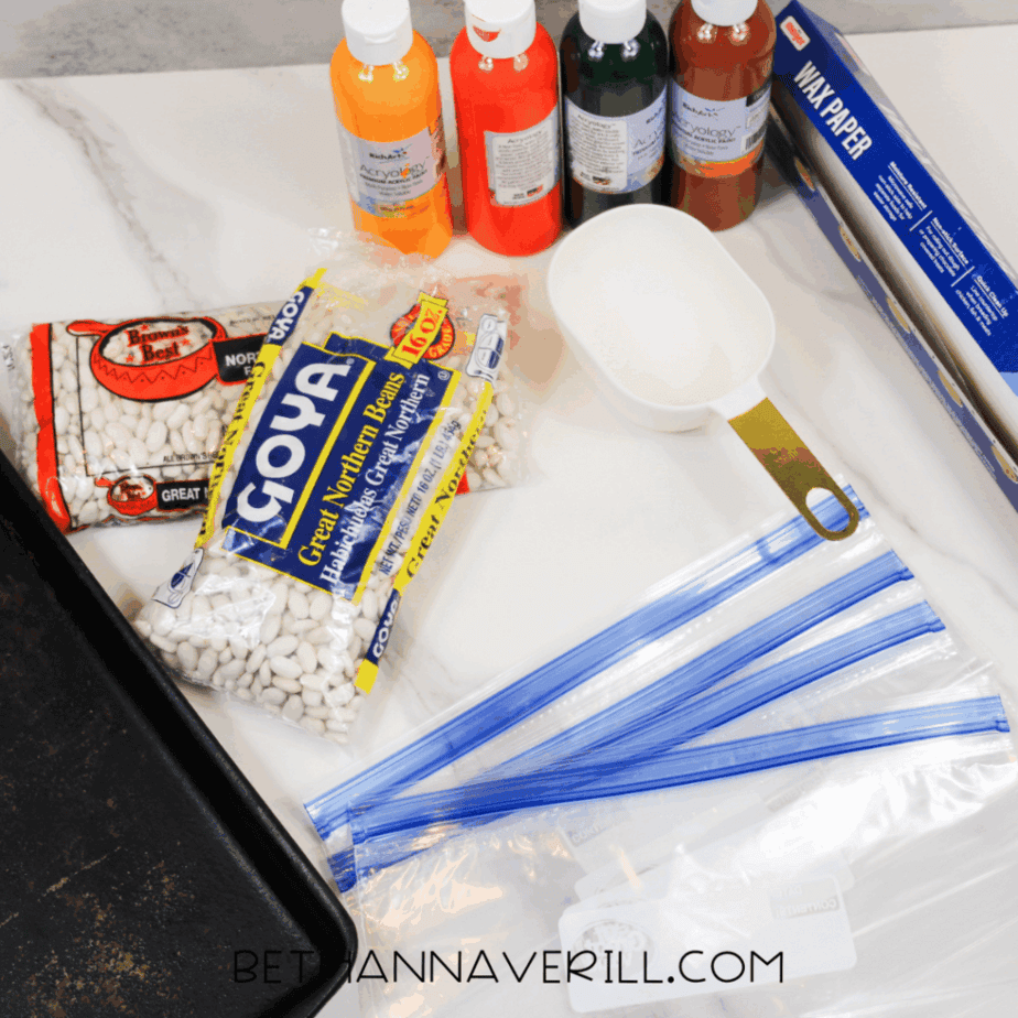Supplies for an autumn sensory bin including dried white beans, acrylic paint in fall colors, zip-top bags, measuring cup, and a plastic sensory tray on a white surface.