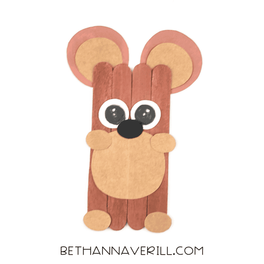 Craft stick bear with layered paper ears, white eye bases, tan muzzle, and black nose, with drawn eyes added to the face.