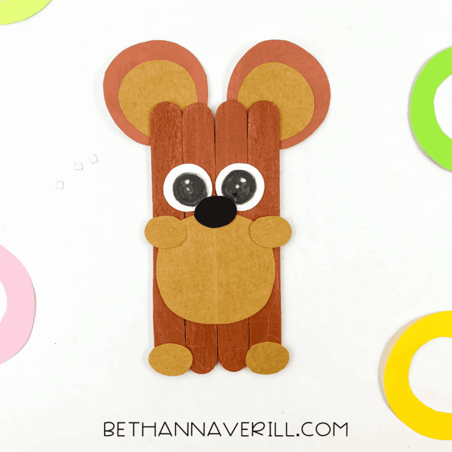 Finished bear popsicle stick craft made from four brown-painted craft sticks with layered paper ears, googly eyes, tan muzzle, black nose, and paper paws on a white background.