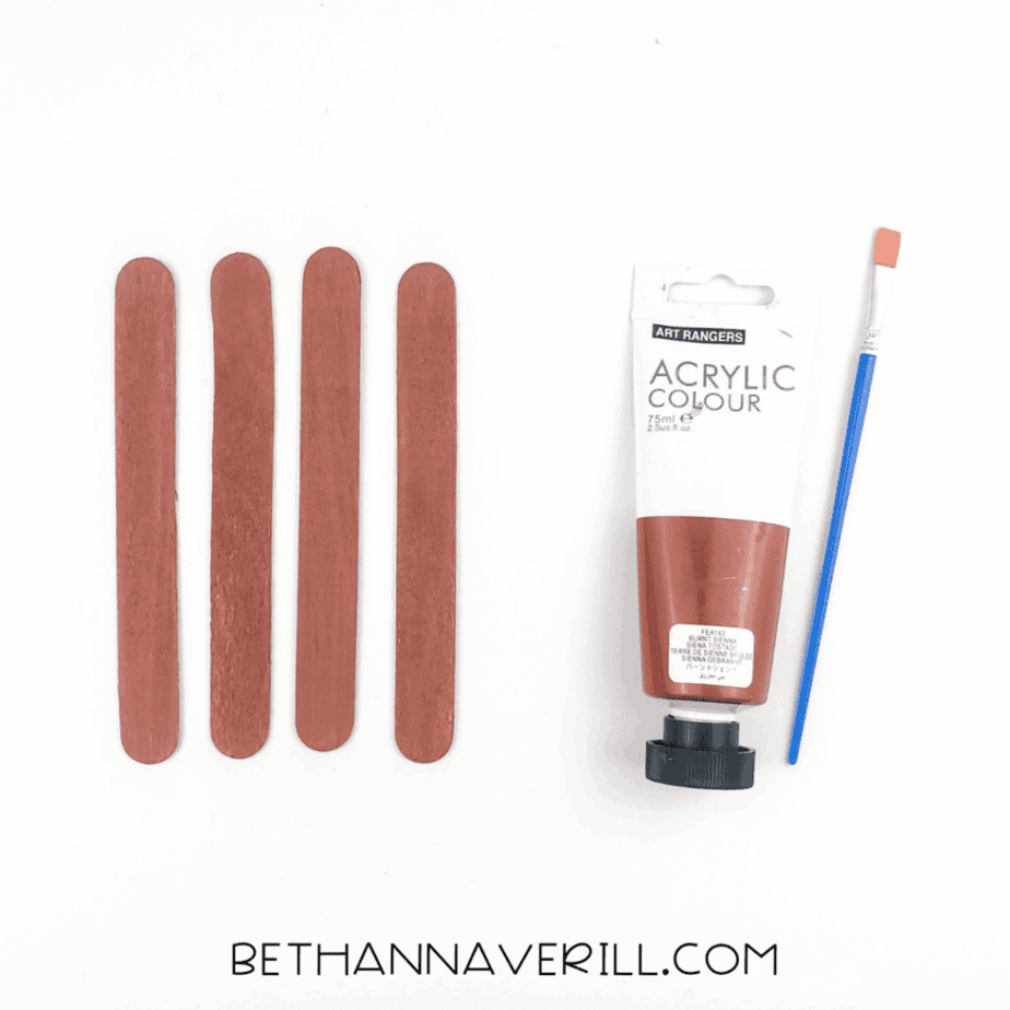 Four jumbo craft sticks painted brown placed side by side next to brown acrylic paint and a paintbrush.