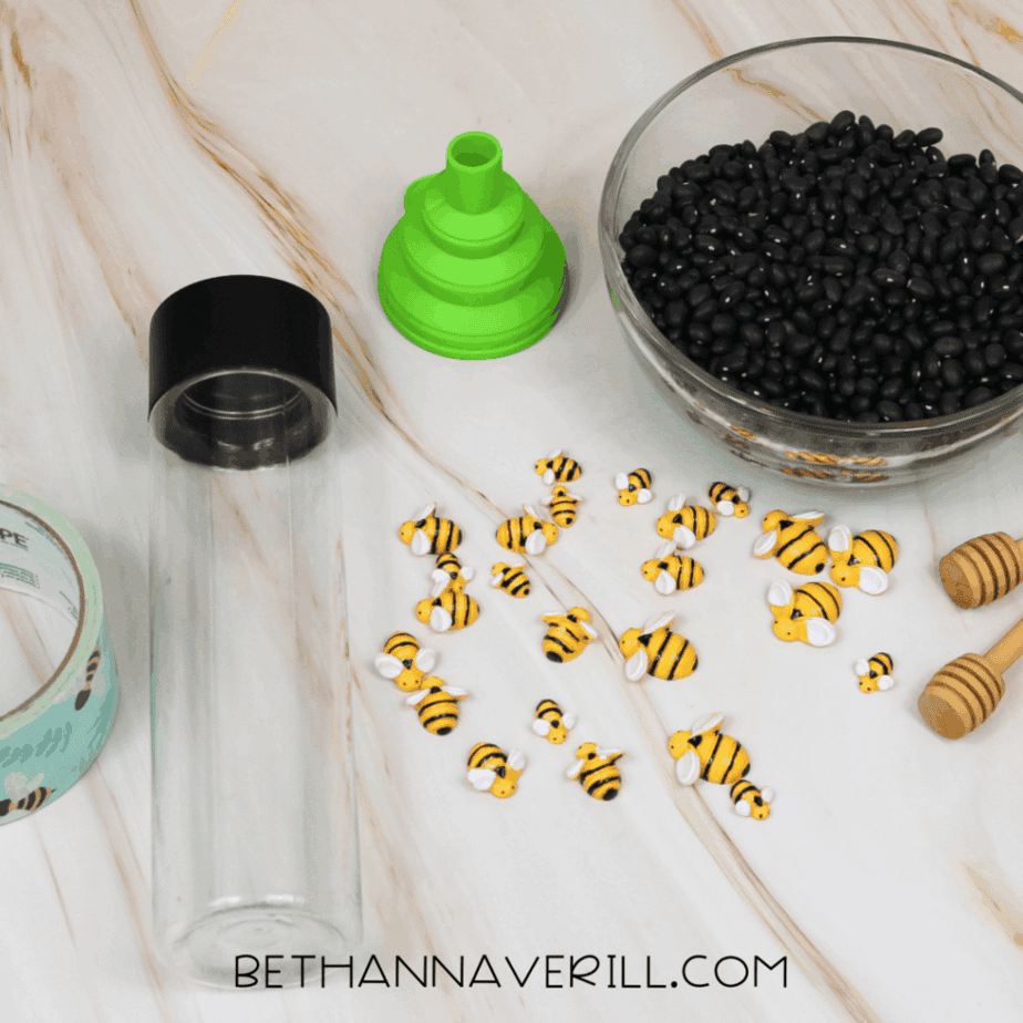 Supplies for a bumble bee sensory bottle including an empty clear bottle, green funnel, black beans in a bowl, small bee figures, honey dippers, and decorative tape.