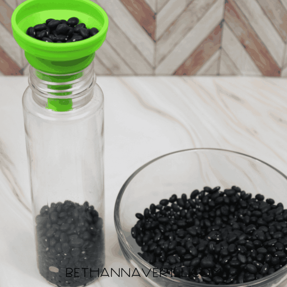 Green funnel placed in the top of a clear bottle while black beans are poured inside.