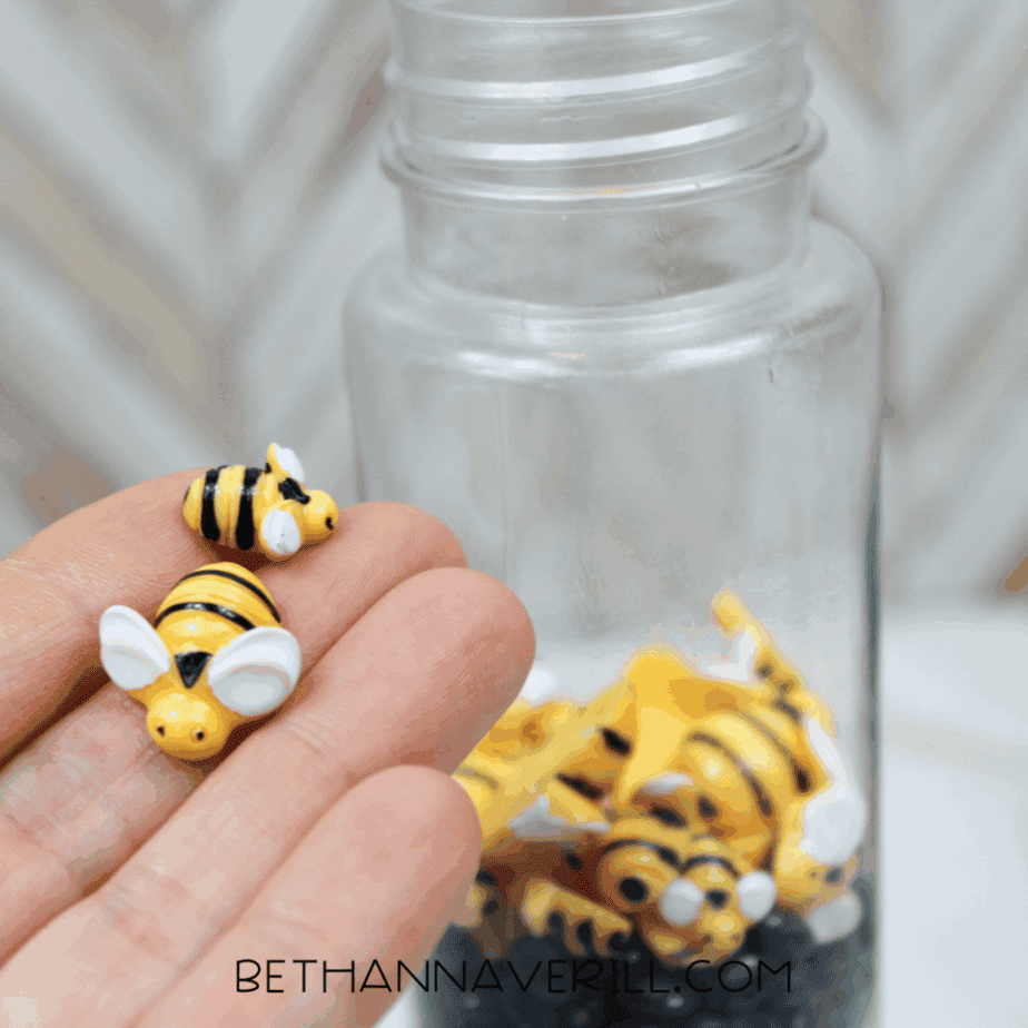 Hand holding two small yellow and black bee figures in front of a partially filled sensory bottle.