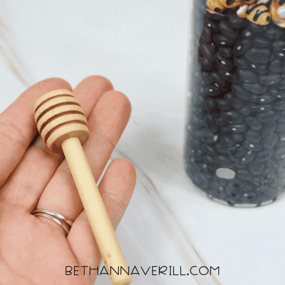 Hand holding a wooden honey dipper next to a sensory bottle filled with black beans and bee figures.