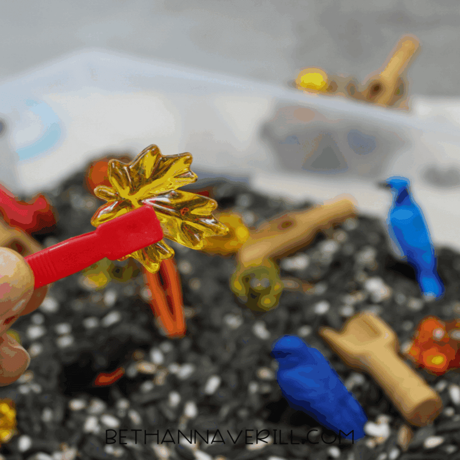 Child’s hand holding red plastic tweezers gripping a yellow translucent leaf gem over a birdseed sensory bin.