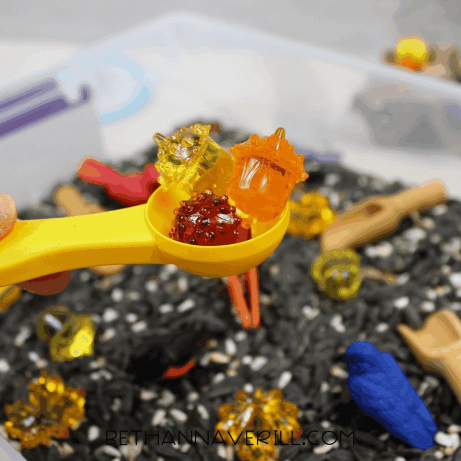 Yellow plastic spoon holding three translucent fall-colored gems above a birdseed sensory bin.