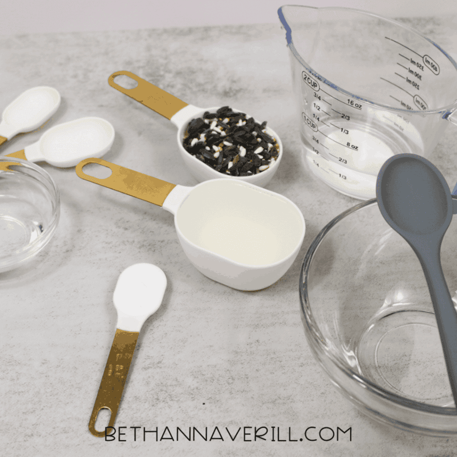 Slime-making supplies arranged on a countertop, including measuring cups, measuring spoons, a glass bowl, a silicone spoon, water, and birdseed.