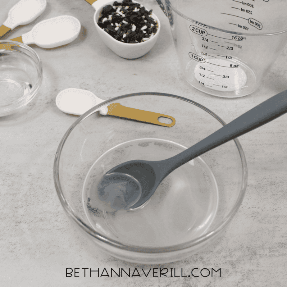 Glass bowl with clear liquid and a gray silicone spoon resting inside, surrounded by measuring tools.