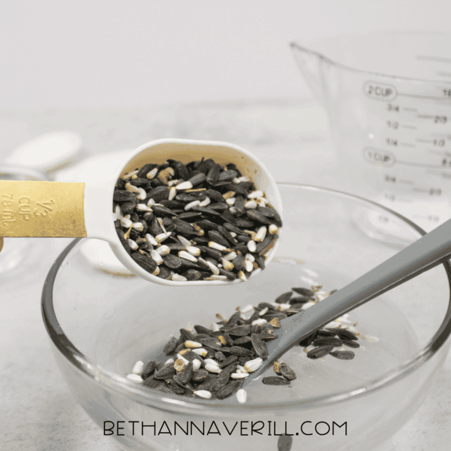 Measuring cup filled with mixed birdseed held above a glass bowl.