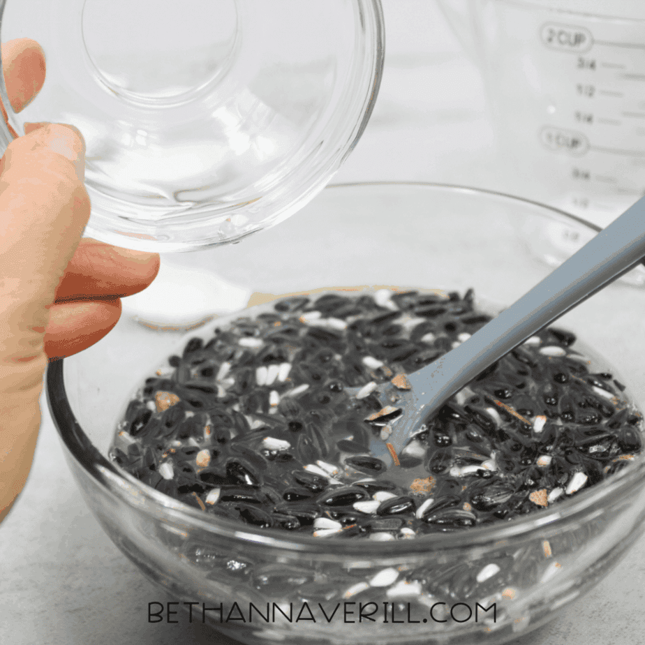 Hand pouring clear liquid into a glass bowl filled with birdseed and liquid mixture.