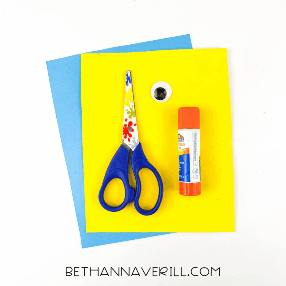 Craft supplies arranged on a white background, including blue and yellow cardstock, child-safe scissors, a glue stick, and a googly eye.
