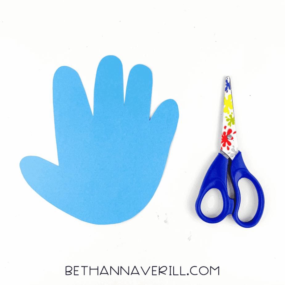 Blue handprint shape cut from cardstock placed next to child-safe scissors on a white surface.