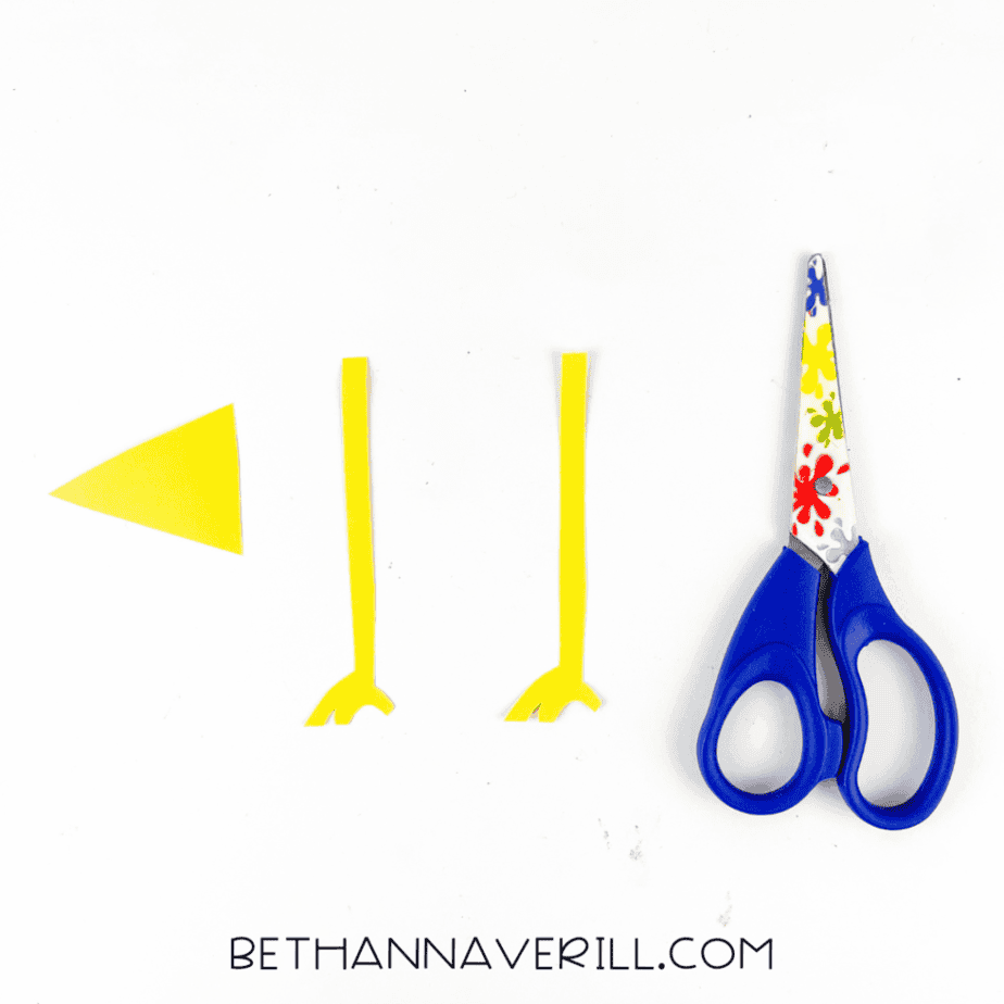 Yellow cardstock pieces cut into a triangle beak shape and two thin leg shapes with feet, arranged beside scissors.