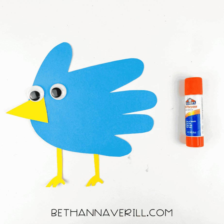 Blue handprint bird craft with googly eyes attached and yellow beak and legs glued in place, shown next to a glue stick.