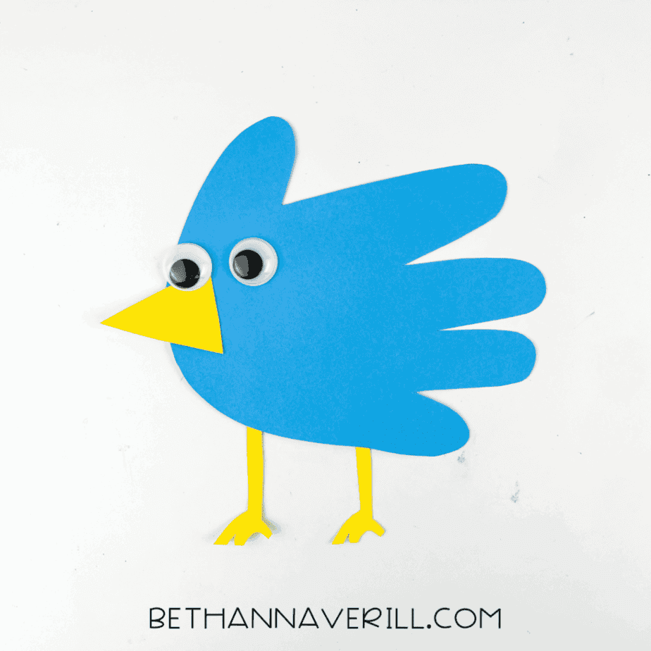 Finished blue bird handprint craft on a white background with googly eyes, yellow beak, and yellow legs.